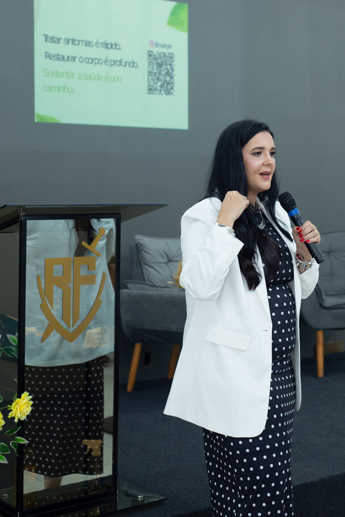 Layla Souza de blazer branco, com microfone na mão, falando durante uma palestra, ao lado do slide projetado